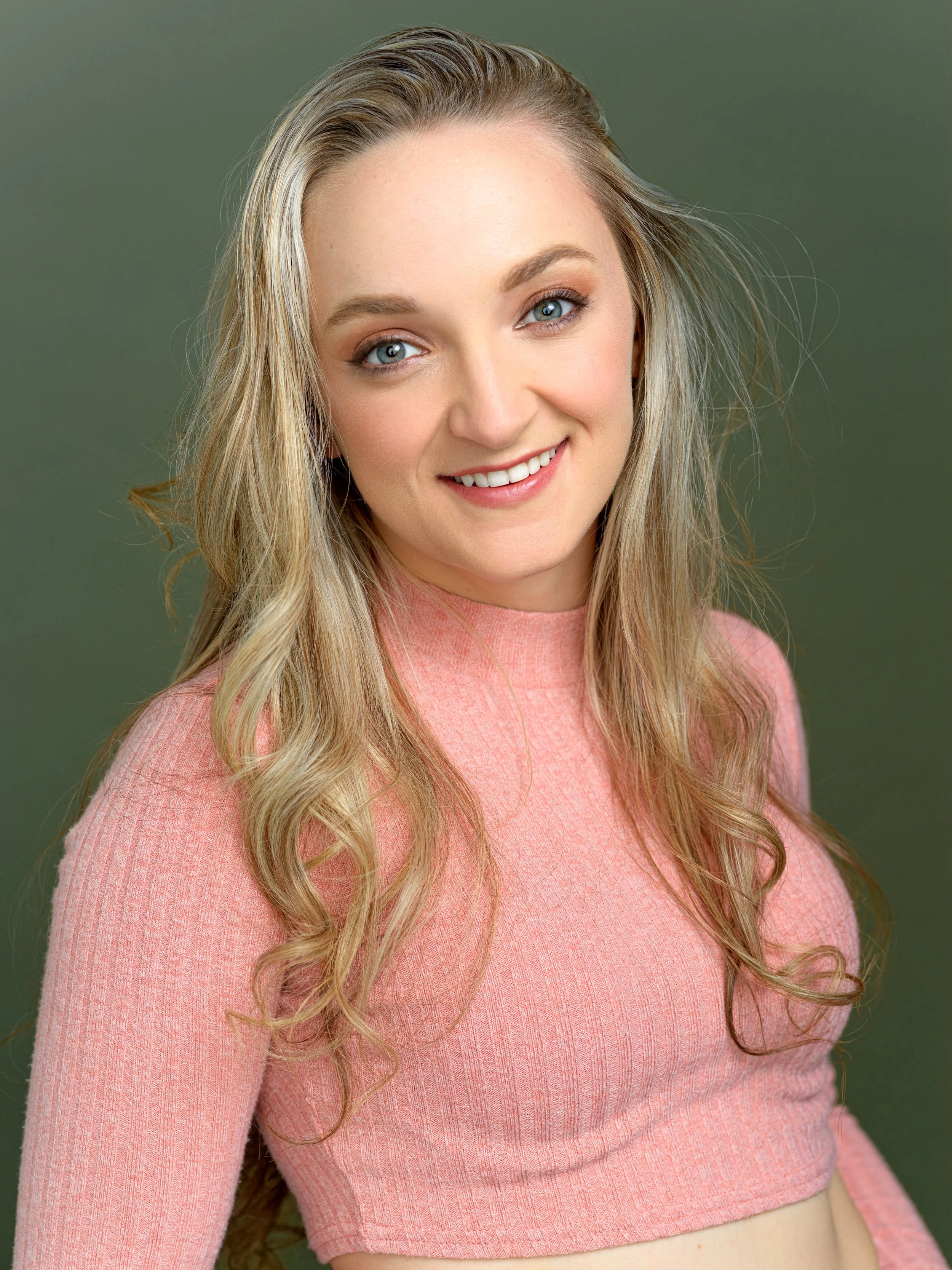 A young woman with long, wavy blonde hair and blue eyes smiling, wearing a pink sweater, against a green background.