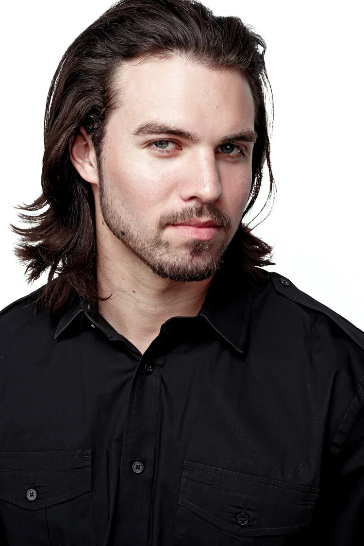 A young man with long brown hair, a beard, and piercing blue eyes, wearing a black button-up shirt, looking directly at the camera with a confident expression.