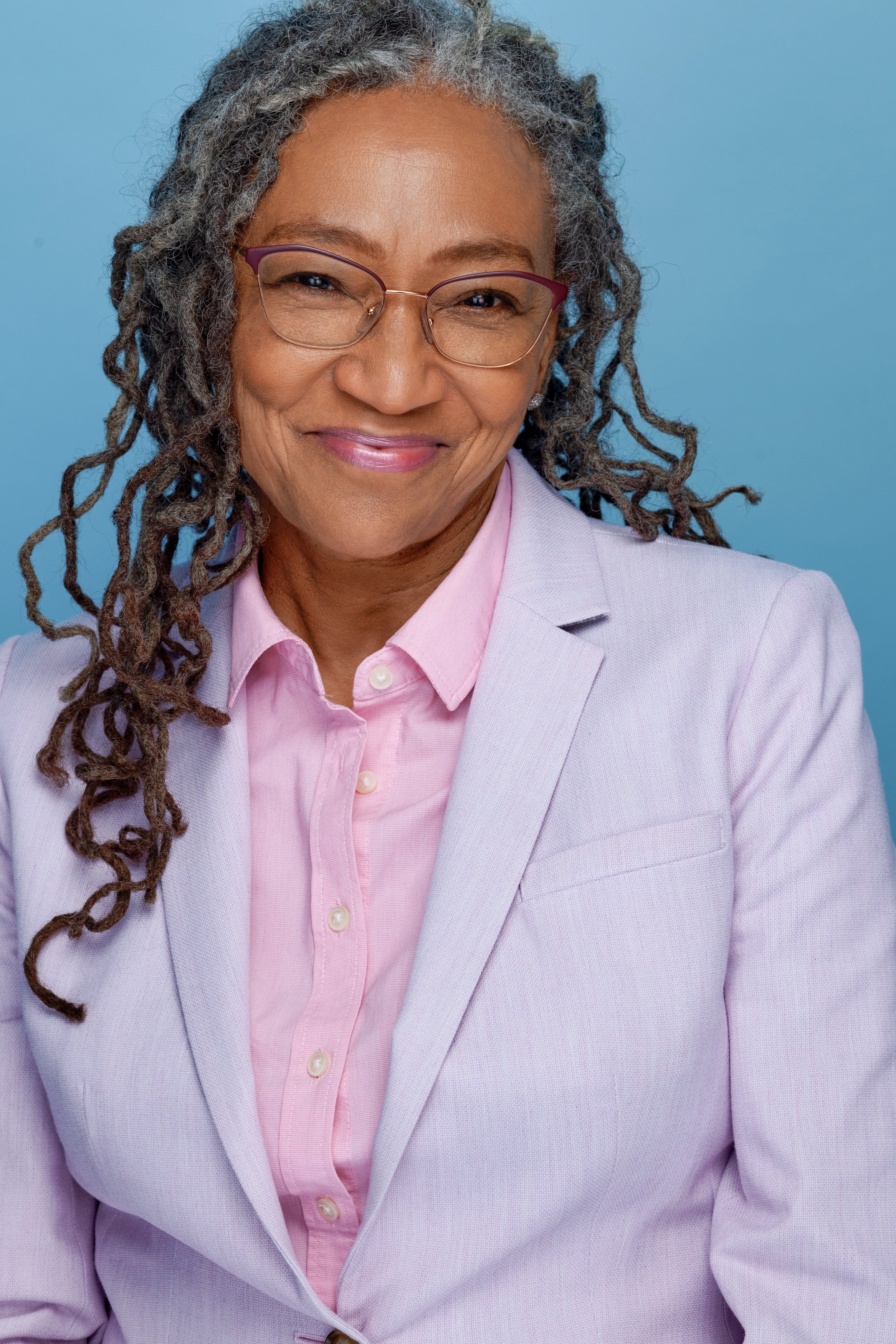 Close-up headshot of a smiling African American woman with gray dreadlocks, wearing pink-rimmed glasses, a lavender blazer, and a pink collared shirt against a blue background.