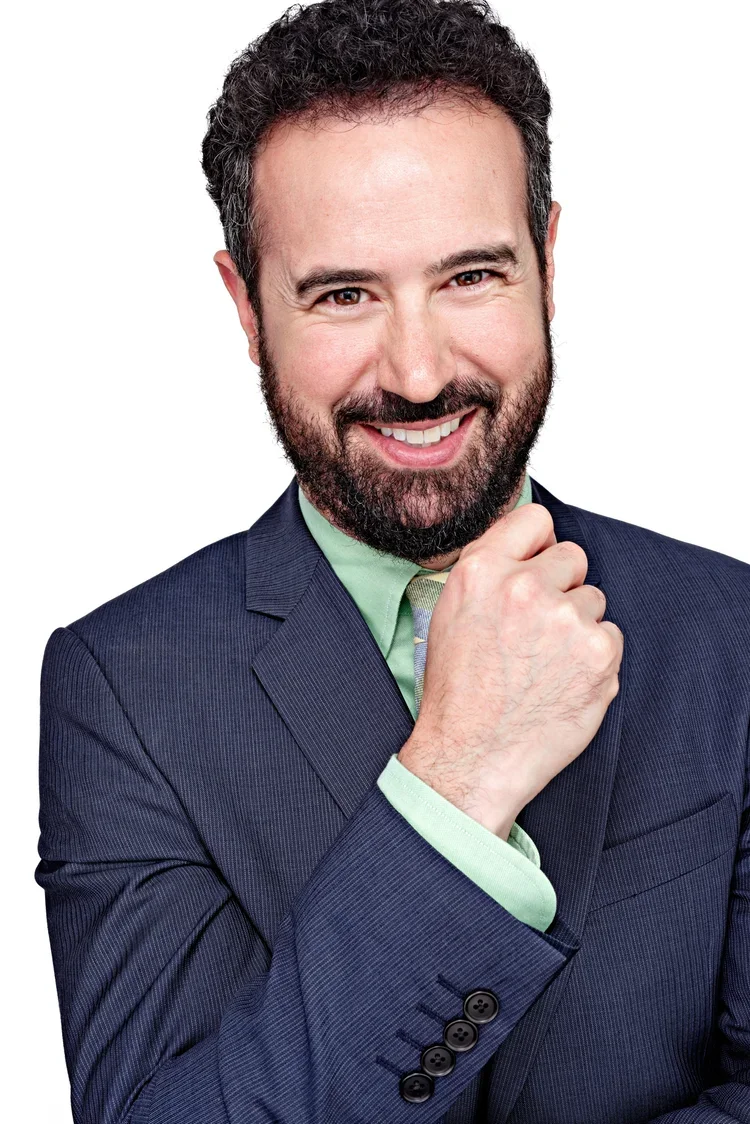 A man with dark curly hair and a beard smiling, wearing a navy blue suit, light green shirt, and light green tie, with his hand resting on his chin.