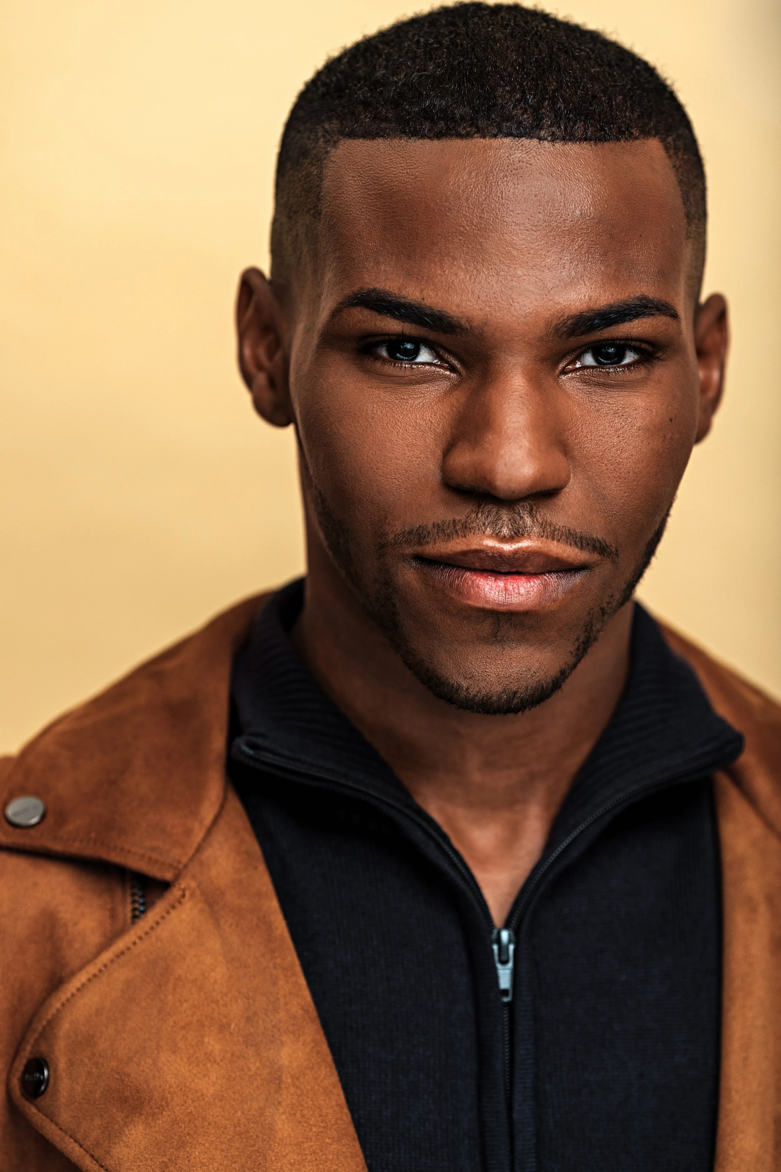 Close-up of a young Black man with short curly hair, wearing a black zip-up jacket and a tan coat, against a beige background.