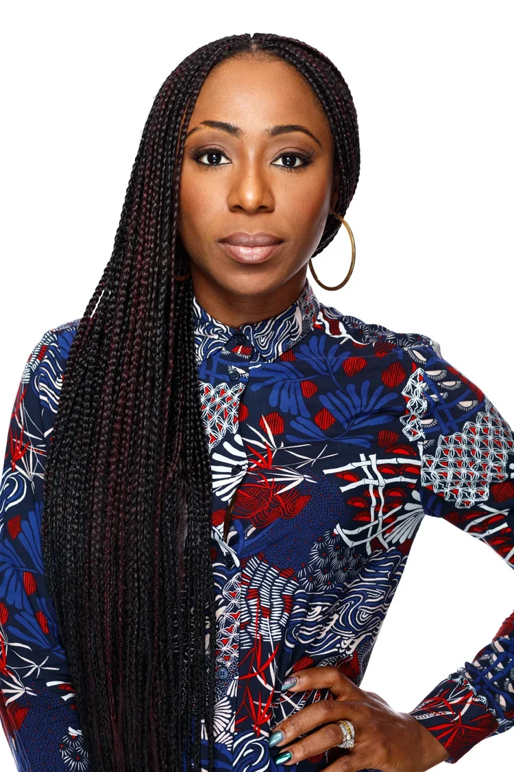 A woman with long braided hair, wearing a colorful patterned shirt in red, blue, white, and black, with one hand on her hip and looking directly at the camera.