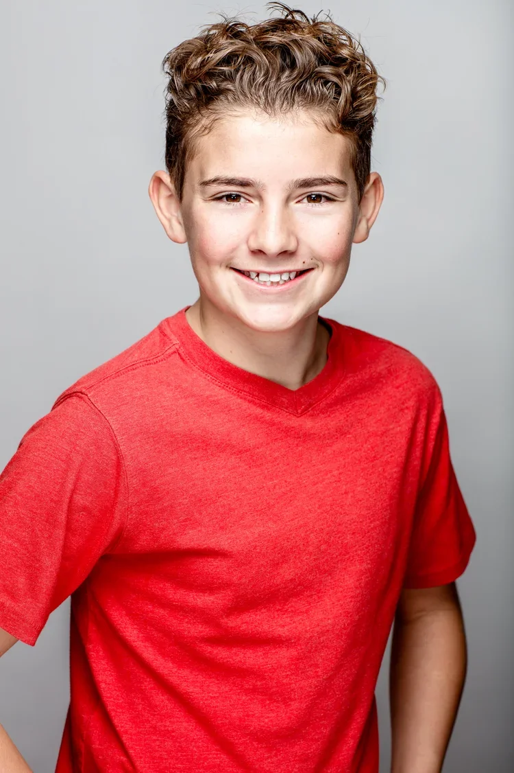 A young boy with curly brown hair wearing a red t-shirt, smiling against a plain gray background.