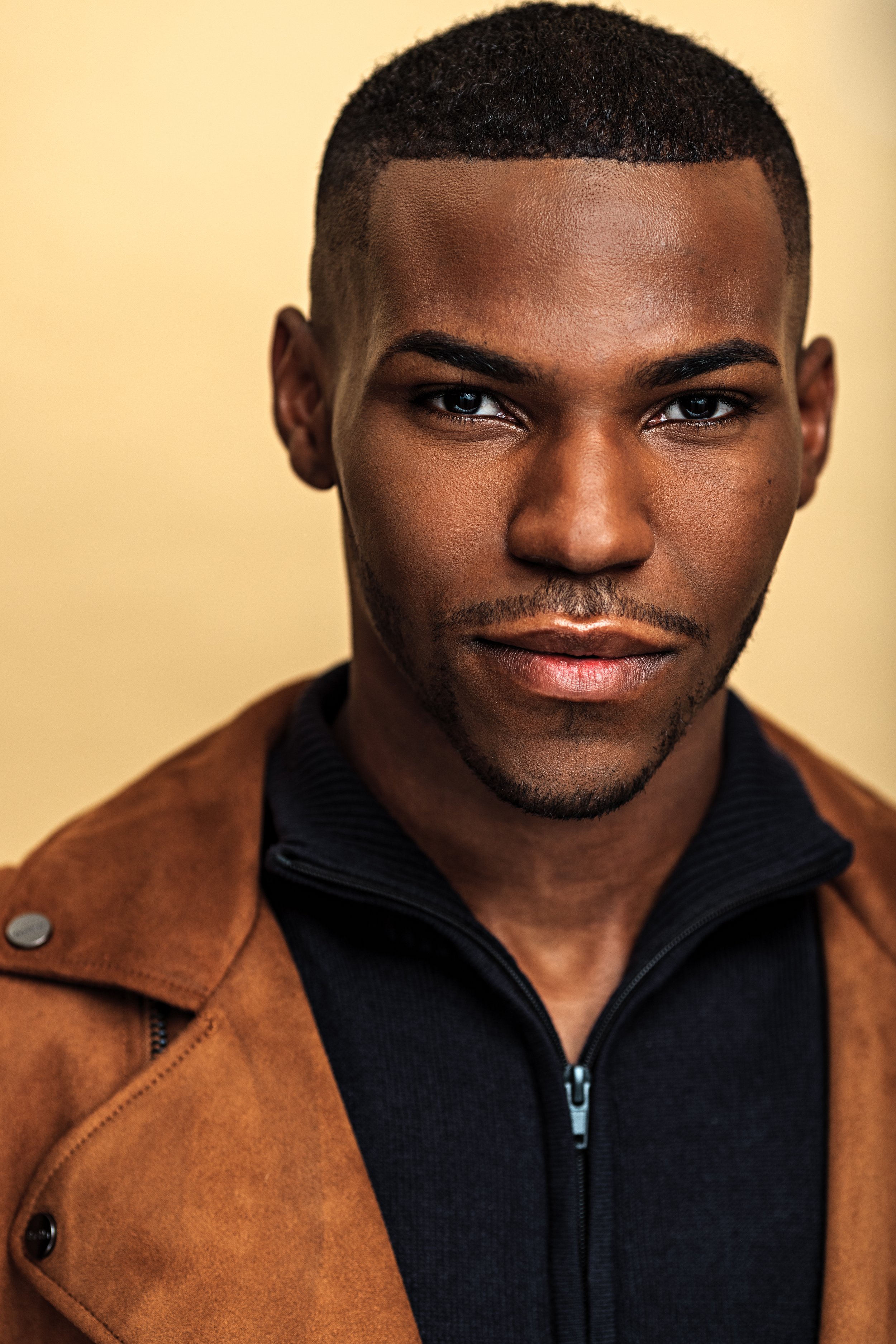Close-up of a young man with dark skin, short black hair, and blue eyes, wearing a brown jacket over a black zip-up hoodie, standing in front of a beige background.