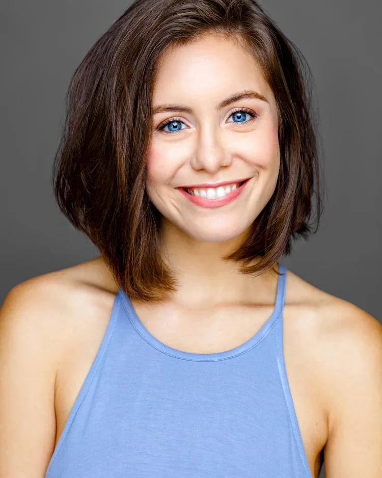 A young woman with shoulder-length brown hair and blue eyes smiling, wearing a light blue sleeveless top against a gray background.