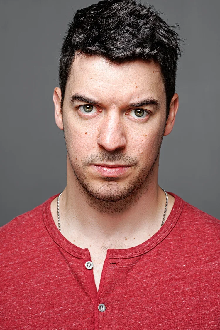 A young man with dark hair and light hazel eyes wearing a red shirt and a silver chain has a neutral expression against a gray background.