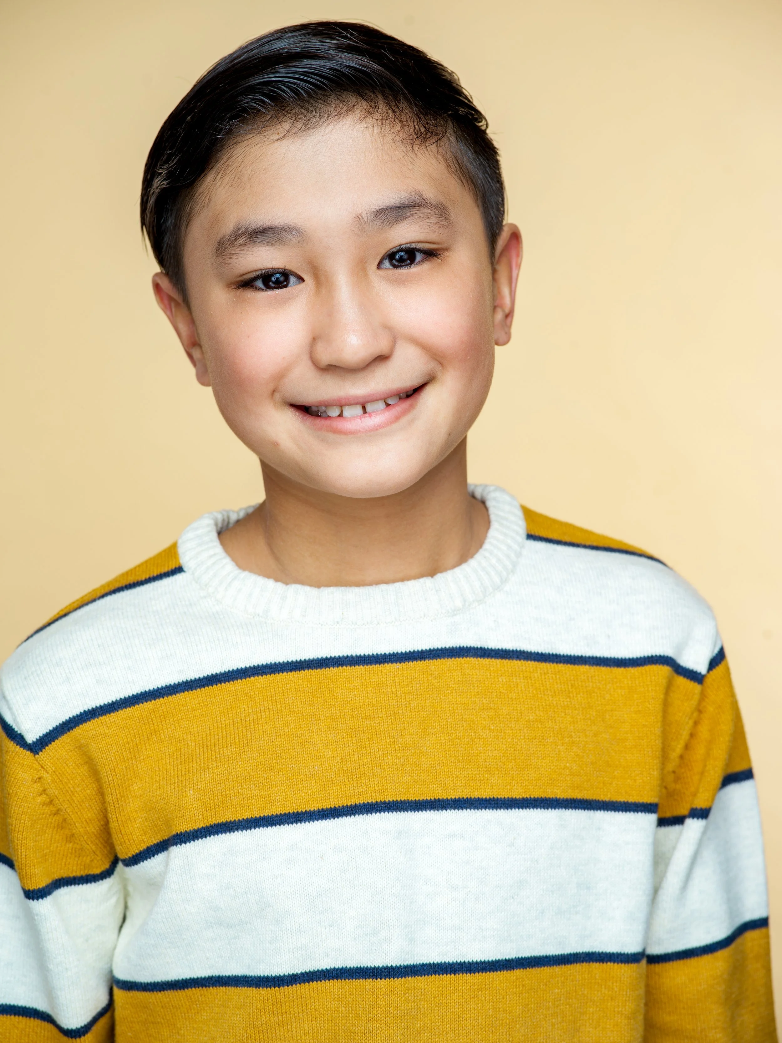 Close-up of a young boy with dark hair, smiling, wearing a yellow, white, and navy striped sweater, against a plain beige background.
