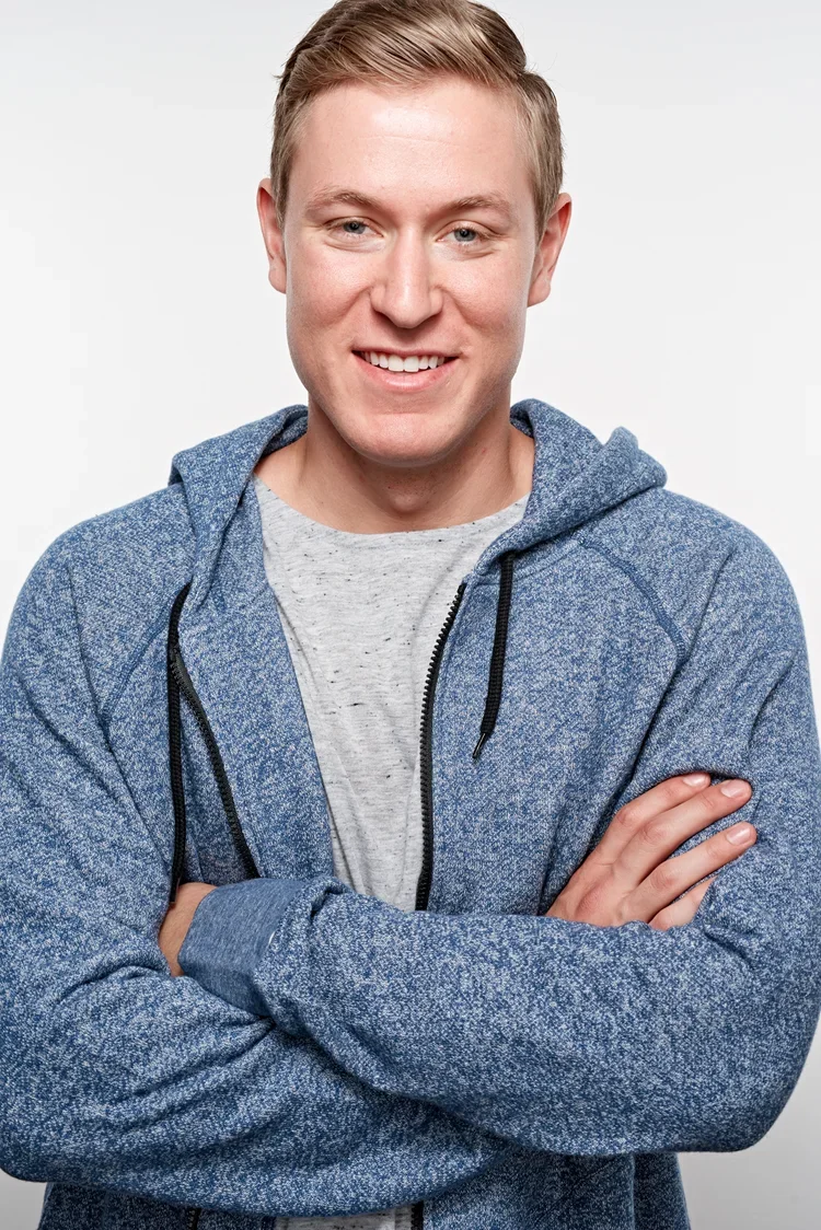 A young man with short blond hair smiling, wearing a gray t-shirt and a blue zip-up hoodie with black drawstrings, standing with arms crossed against a plain gray background.