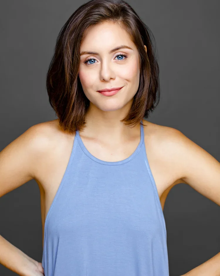 Young woman with shoulder-length brown hair and blue eyes, wearing a light blue tank top, smiling confidently against a dark gray background.
