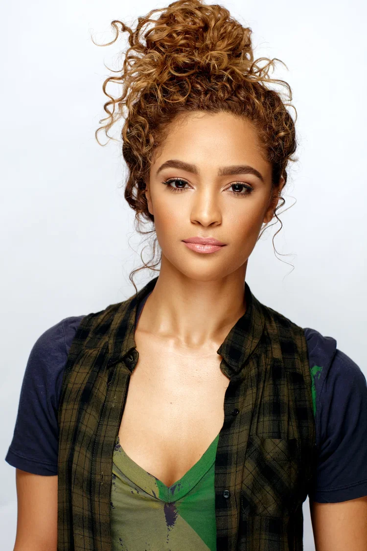 A young woman with curly auburn hair styled in a high bun, wearing a dark plaid vest over a green top, posing against a plain white background.