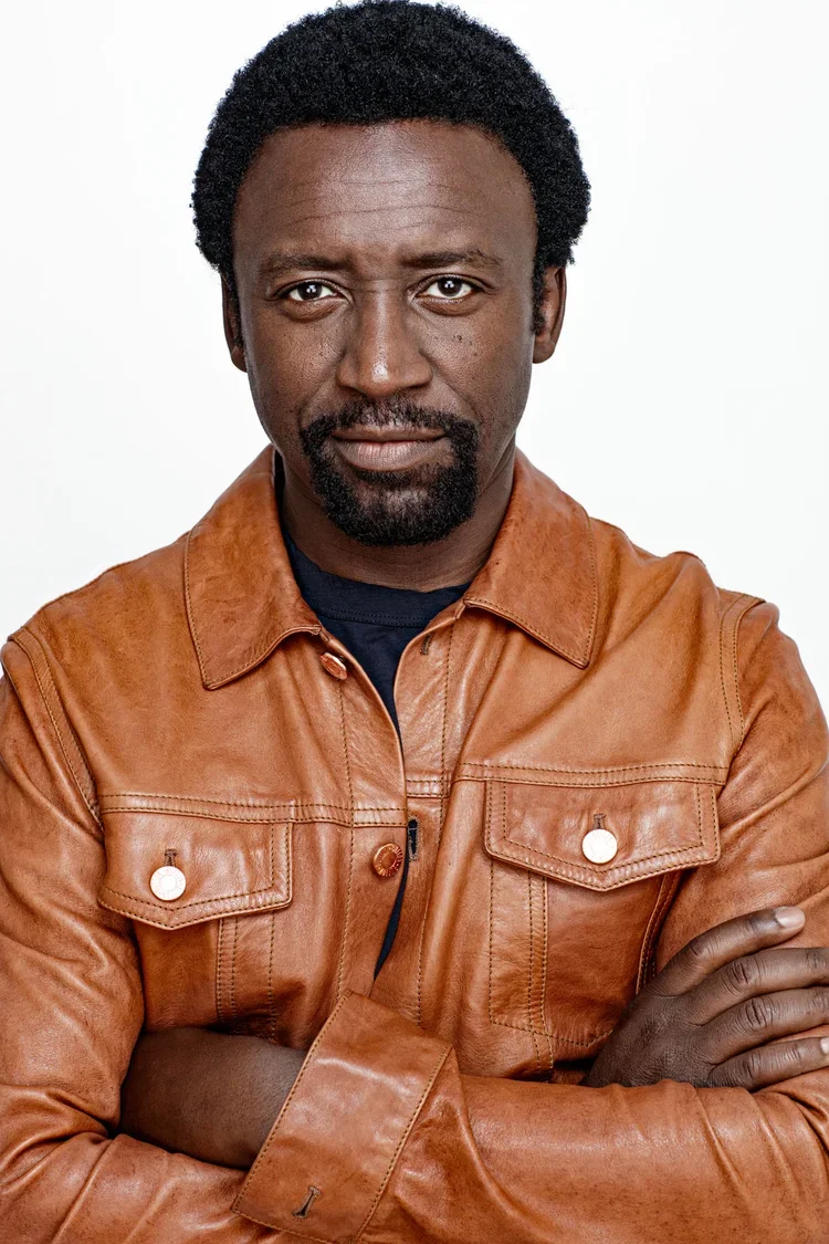 A man with dark skin and a serious expression, wearing a brown leather jacket and crossing his arms, standing against a white background.