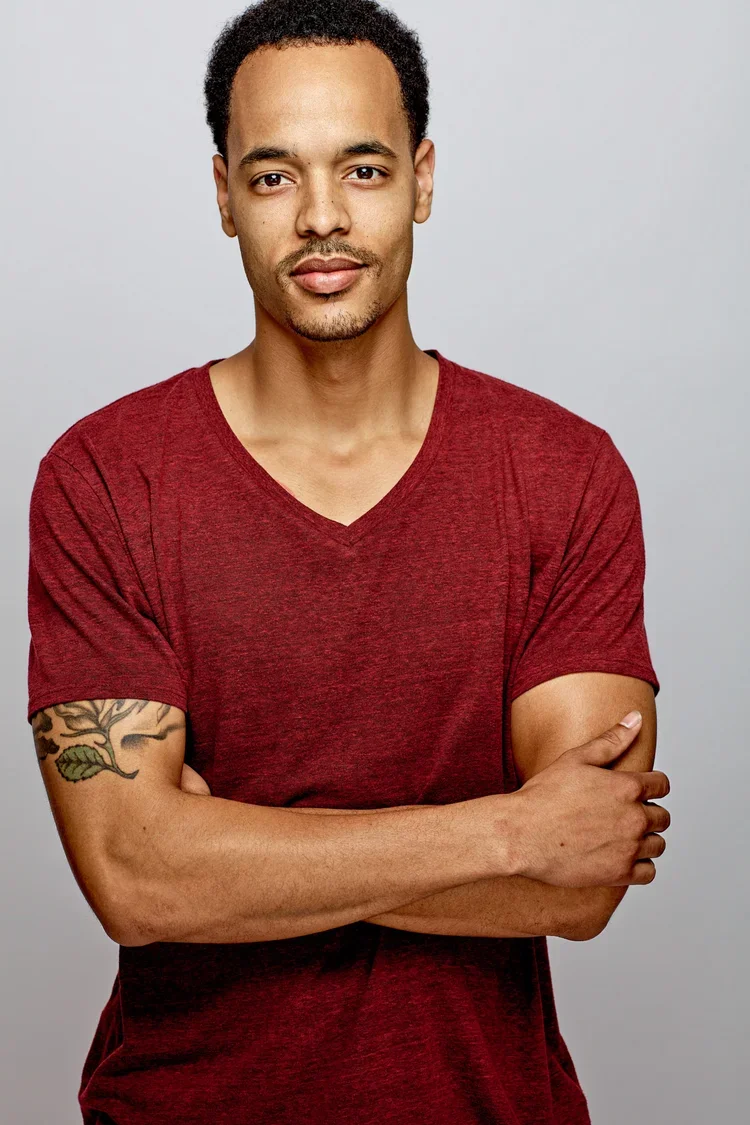 A man with short curly hair, a slight beard, wearing a red V-neck t-shirt, crossing his arms, standing against a plain light gray background.