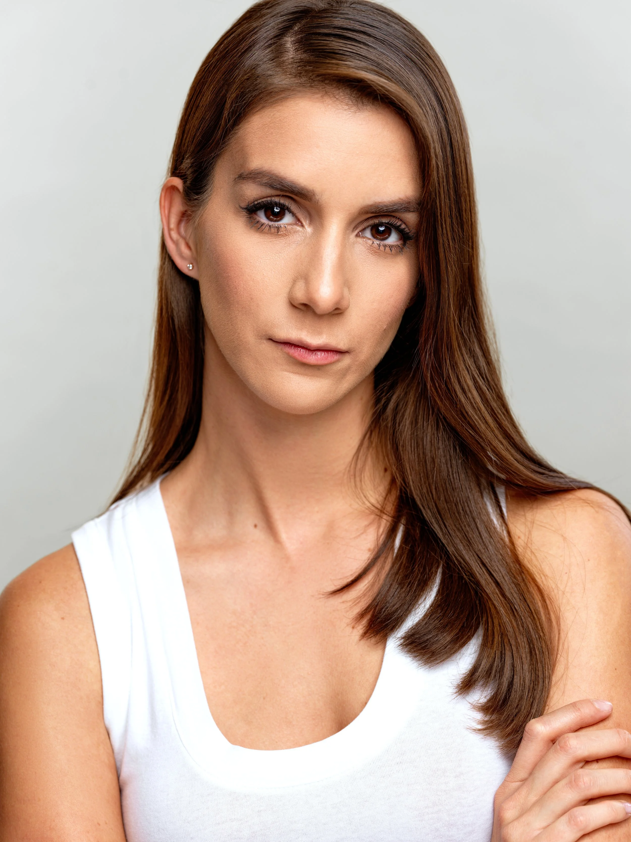 Close-up of a woman with brown hair wearing a white top, with her face turned slightly to the side, showing her brown eyes, earrings, and a gentle expression.