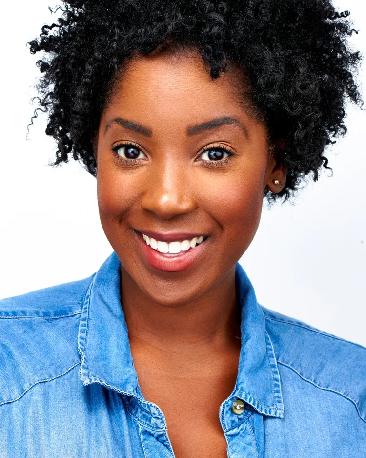 Close-up portrait of a smiling Black woman with curly black hair, wearing a denim shirt against a plain white background.