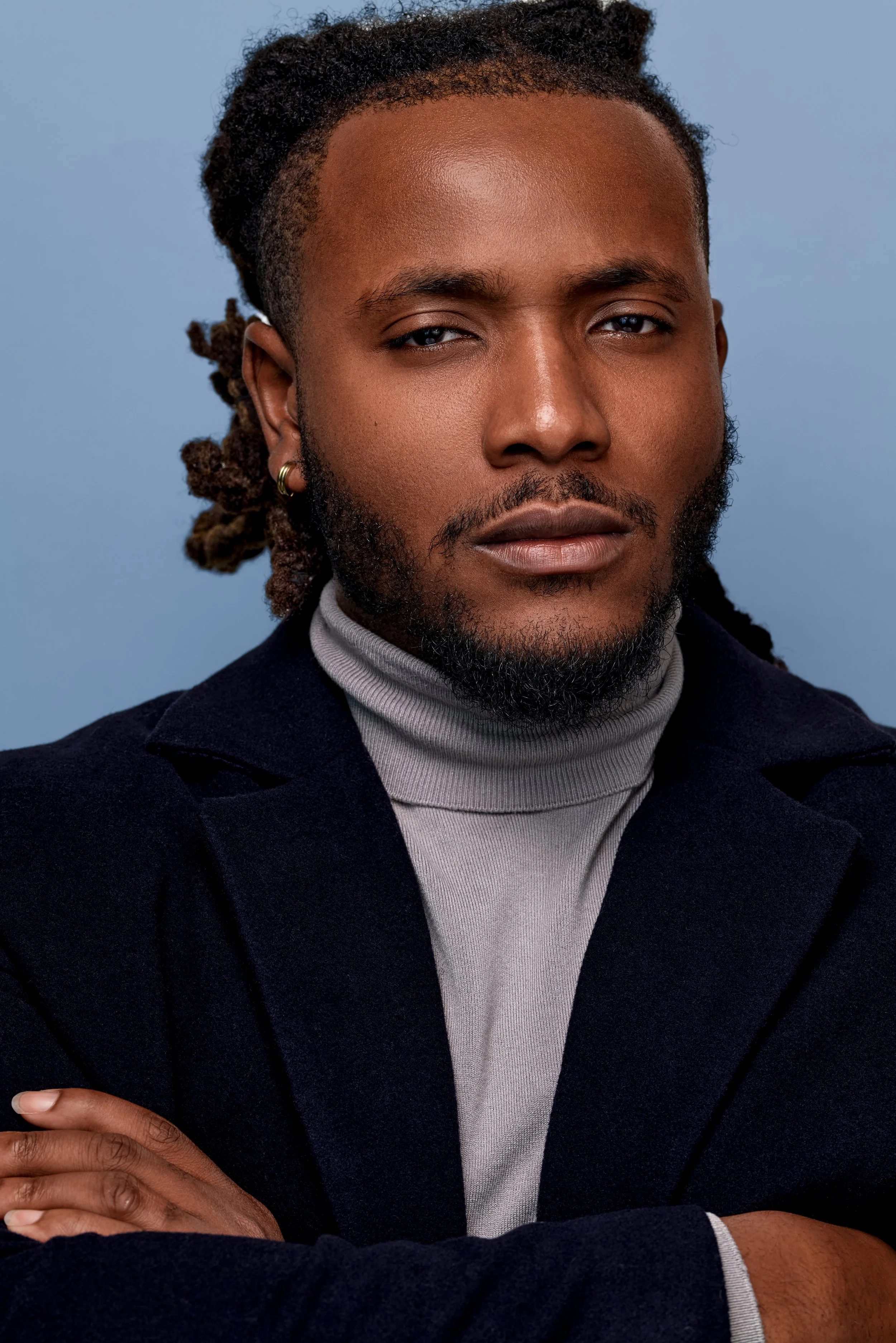 Close-up of a man with medium brown skin, dreadlocked hair, and a beard, wearing a gray turtleneck and a dark blazer, against a blue background.