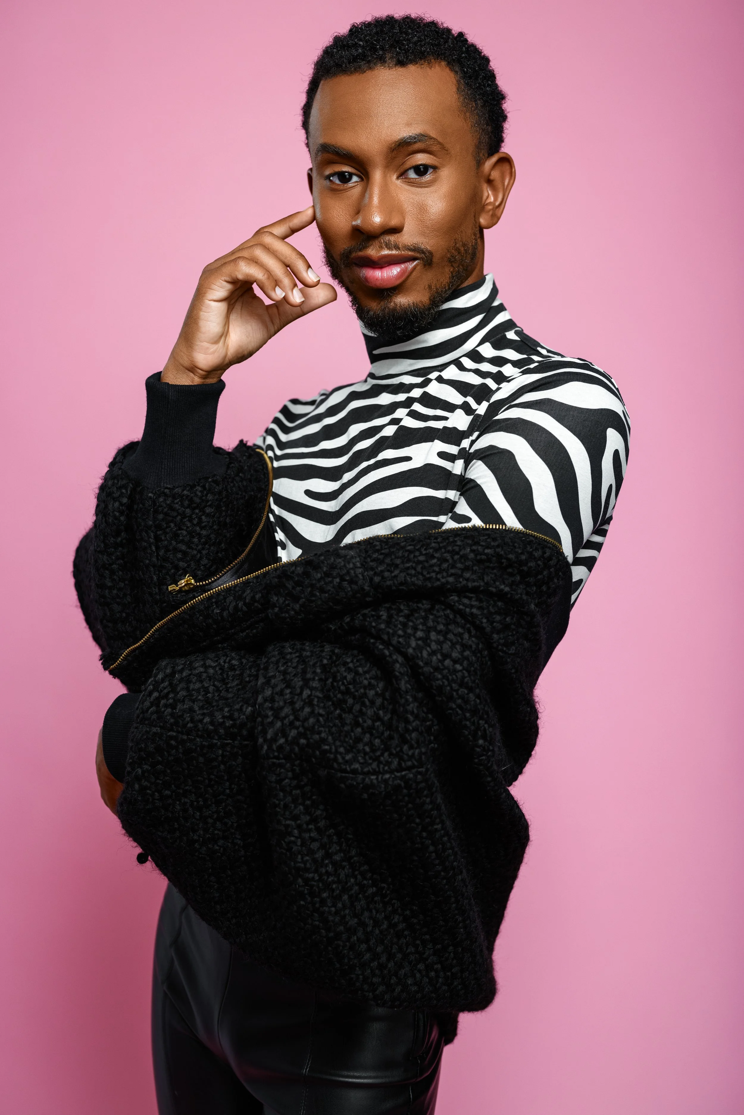 A young man with a confident expression, wearing a black and white striped turtleneck and a black textured jacket, standing against a pink background.