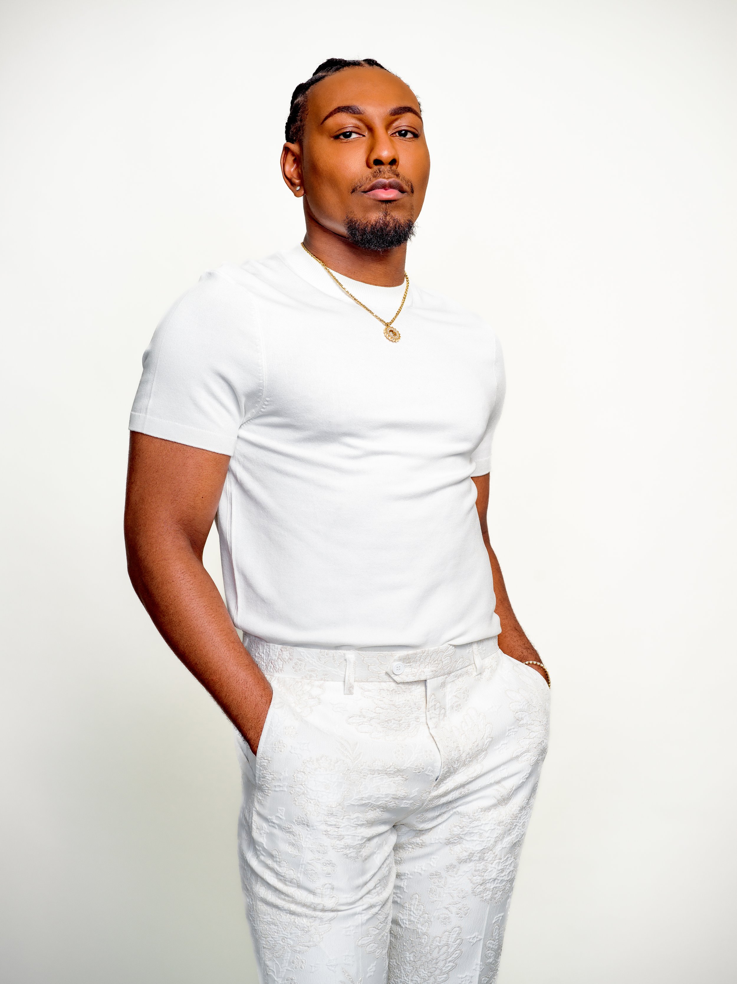 Portrait of a confident man with short dreadlocks, a goatee, wearing a white t-shirt, white patterned pants, gold chain necklace, and a bracelet, standing against a plain white background.