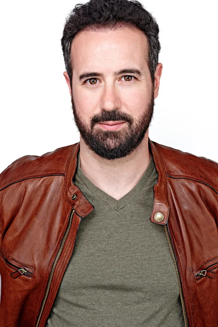 A man with curly dark hair and a beard wearing a green V-neck shirt and a brown leather jacket, looking at the camera with a neutral expression.