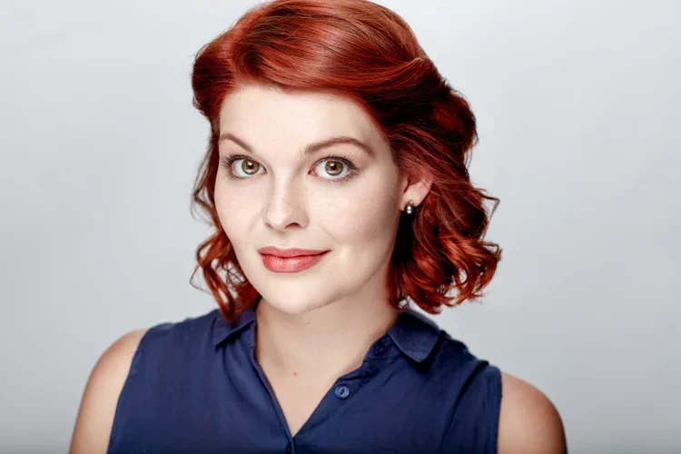 A woman with short, curly red hair and fair skin, wearing a sleeveless navy blue top, looking at the camera with a slight smile.