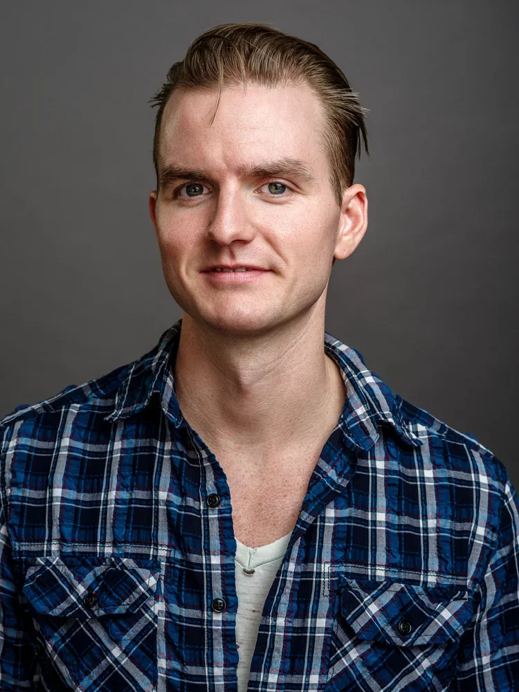 Portrait of a young man with light skin and light brown hair wearing a blue plaid shirt against a gray background.