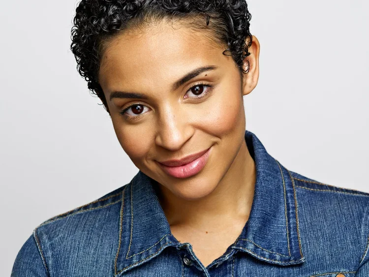 Close-up portrait of a young woman with short curly hair, smiling, wearing a denim jacket against a plain light background.