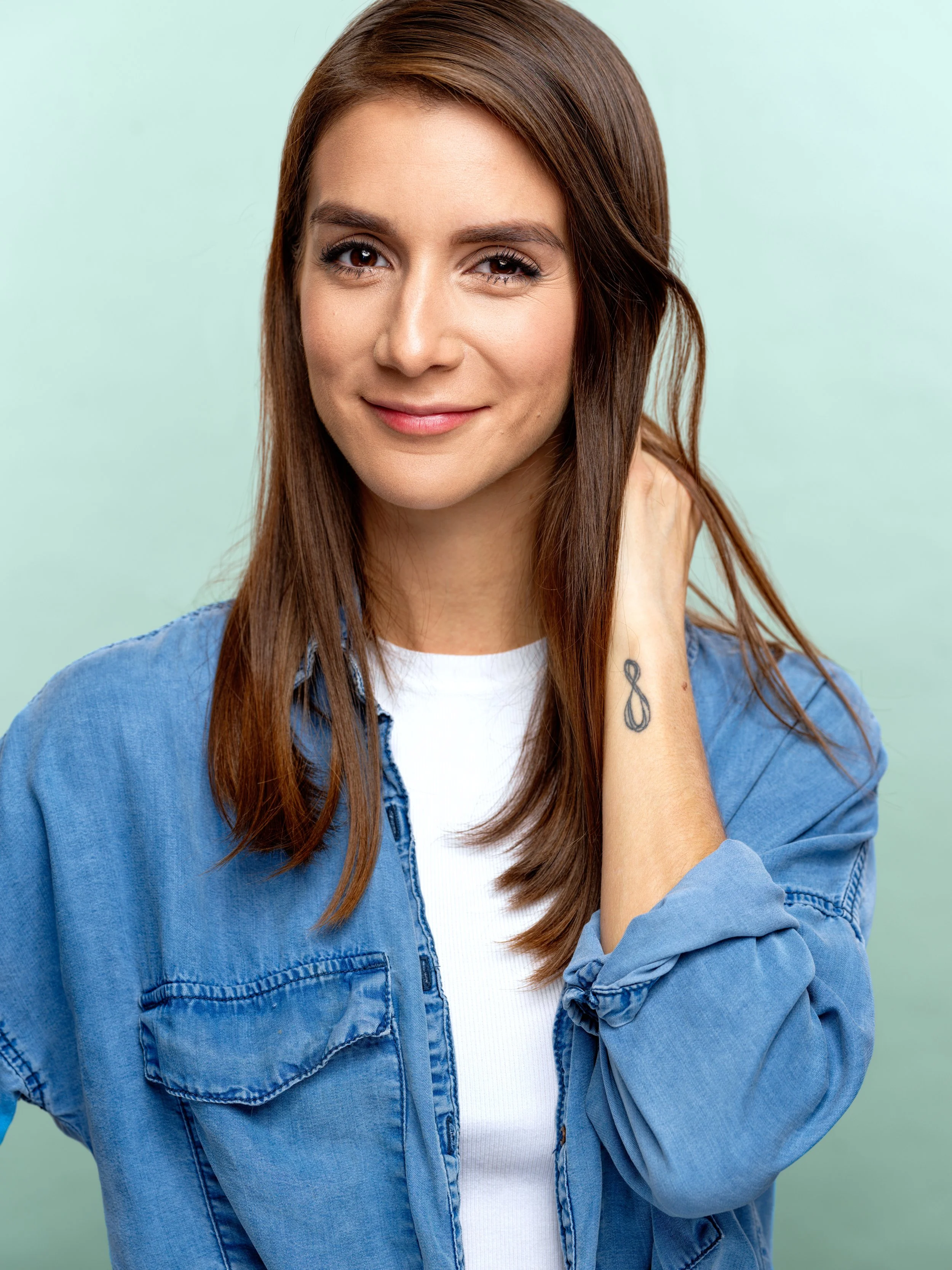 A young woman with long brown hair pulled to one side, wearing a white shirt and blue denim jacket, smiling slightly with one eye winking, against a light green background.