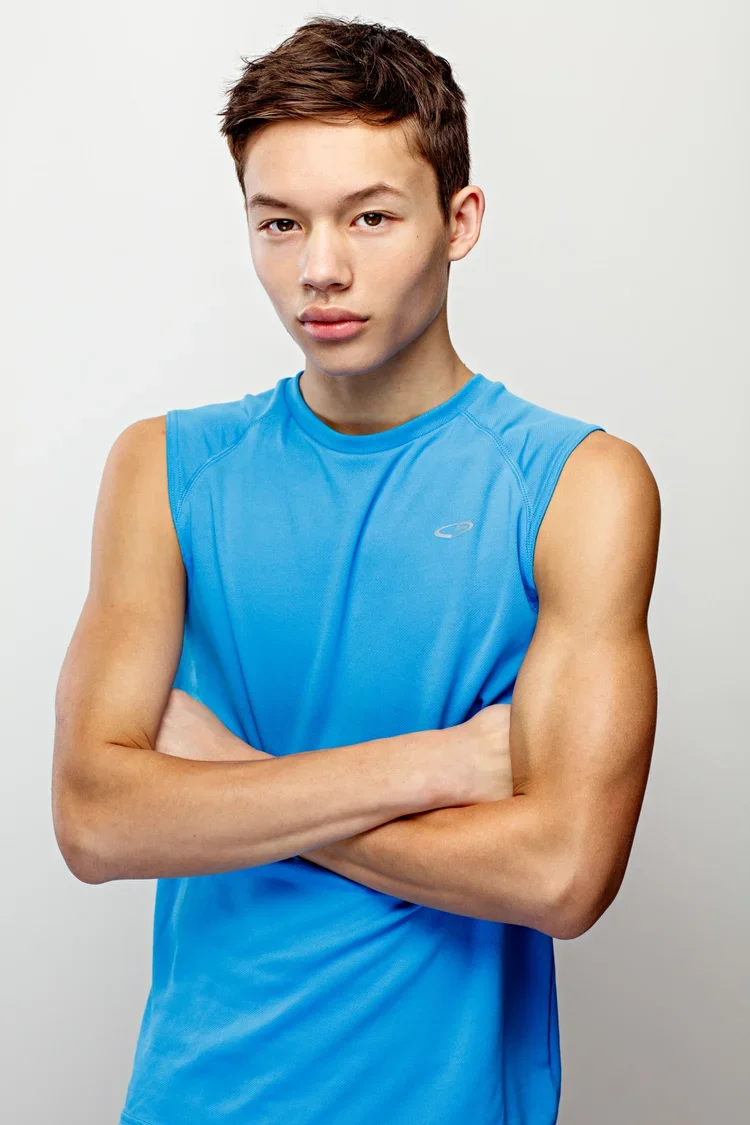 Young man wearing a blue sports tank top with his arms crossed, standing against a plain background.