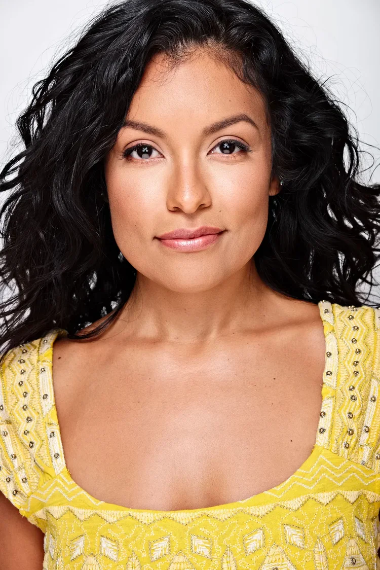 Close-up portrait of a young woman with wavy black hair, wearing a yellow embroidered top, smiling softly with natural makeup, against a plain light gray background.