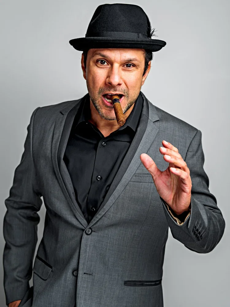 Man wearing a gray suit and black shirt, with a black fedora, holding a cigar in his mouth, gesturing with his right hand against a gray background.