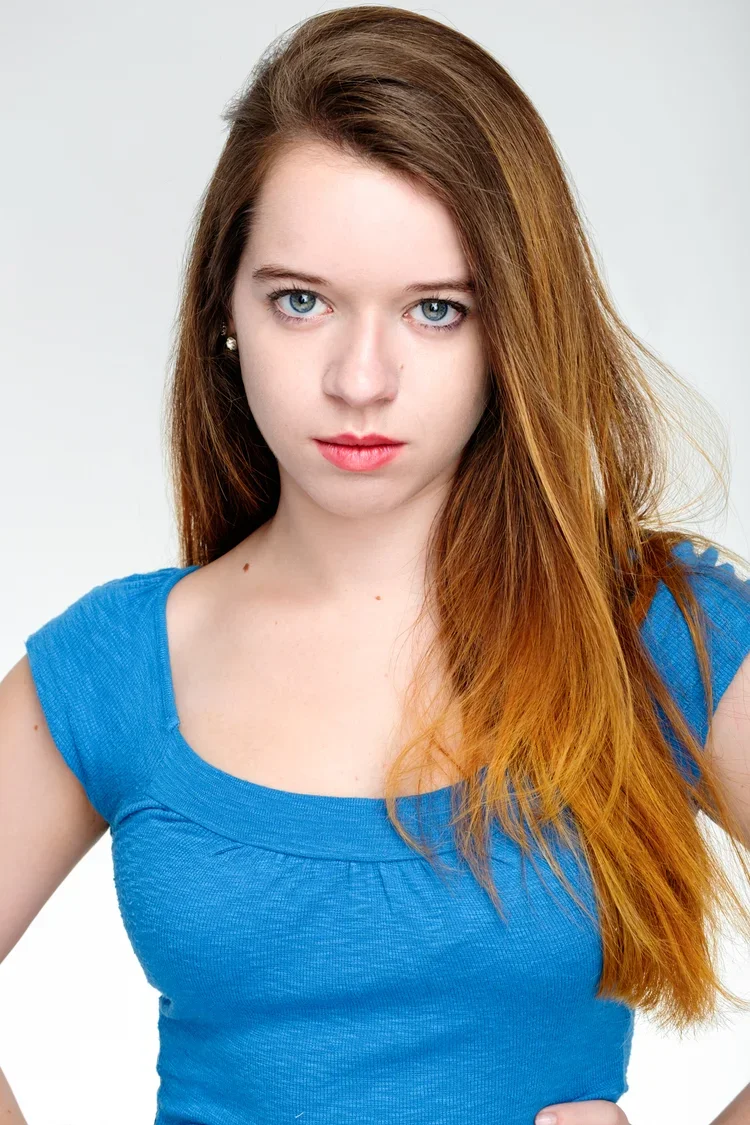 Close-up of a young woman with red hair, blue eyes, wearing a blue top, against a plain white background.