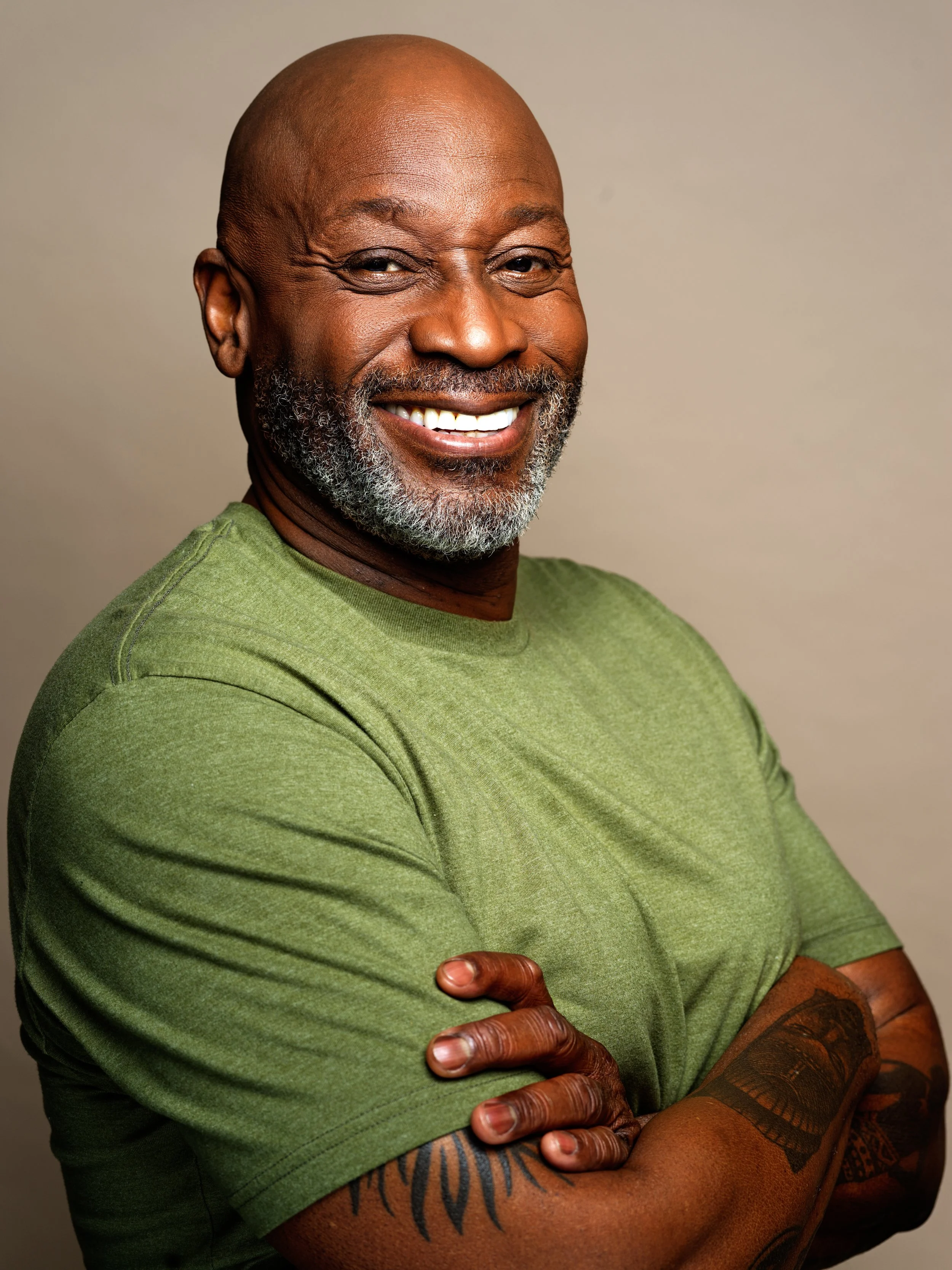 A smiling, bald African-American man with a beard, wearing a green t-shirt, showing tattoos on his arm, with arms crossed and pointing with his finger.