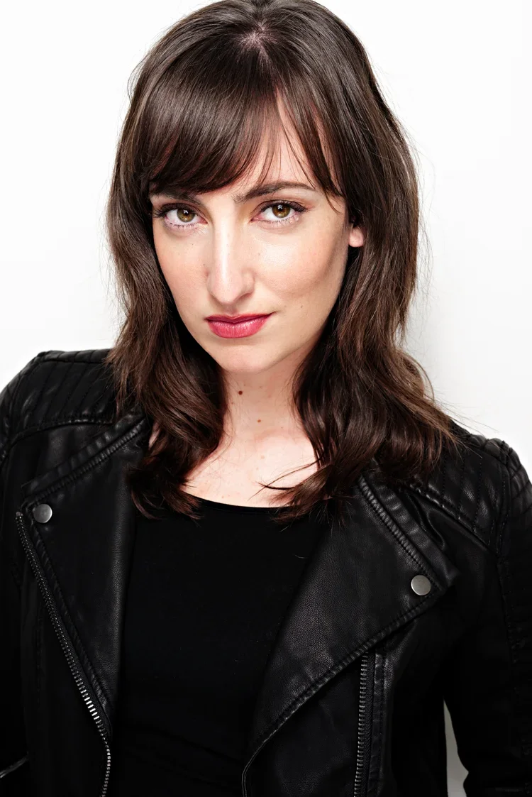 A woman with shoulder-length brown hair, wearing a black leather jacket and black top, looking directly at the camera against a plain white background.