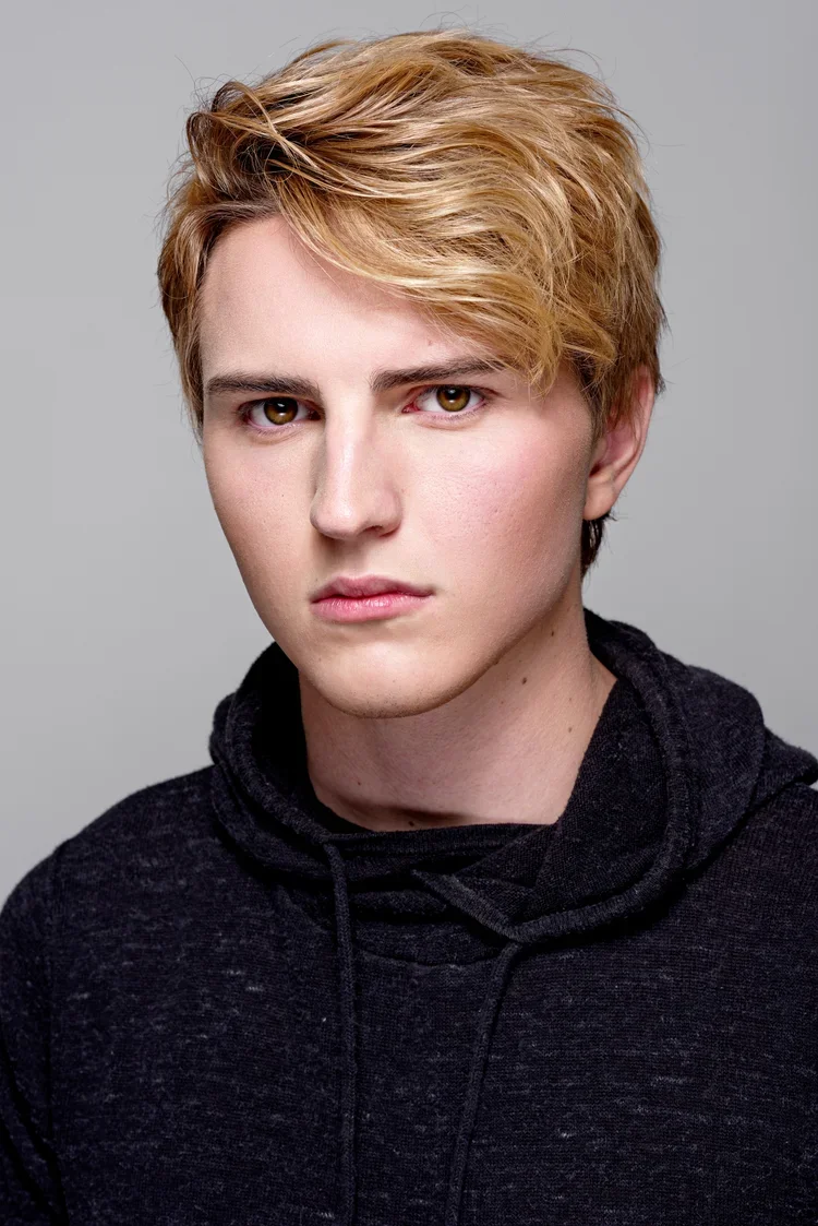 A young man with light skin, blonde hair, and brown eyes, wearing a black hoodie, facing the camera against a gray background.