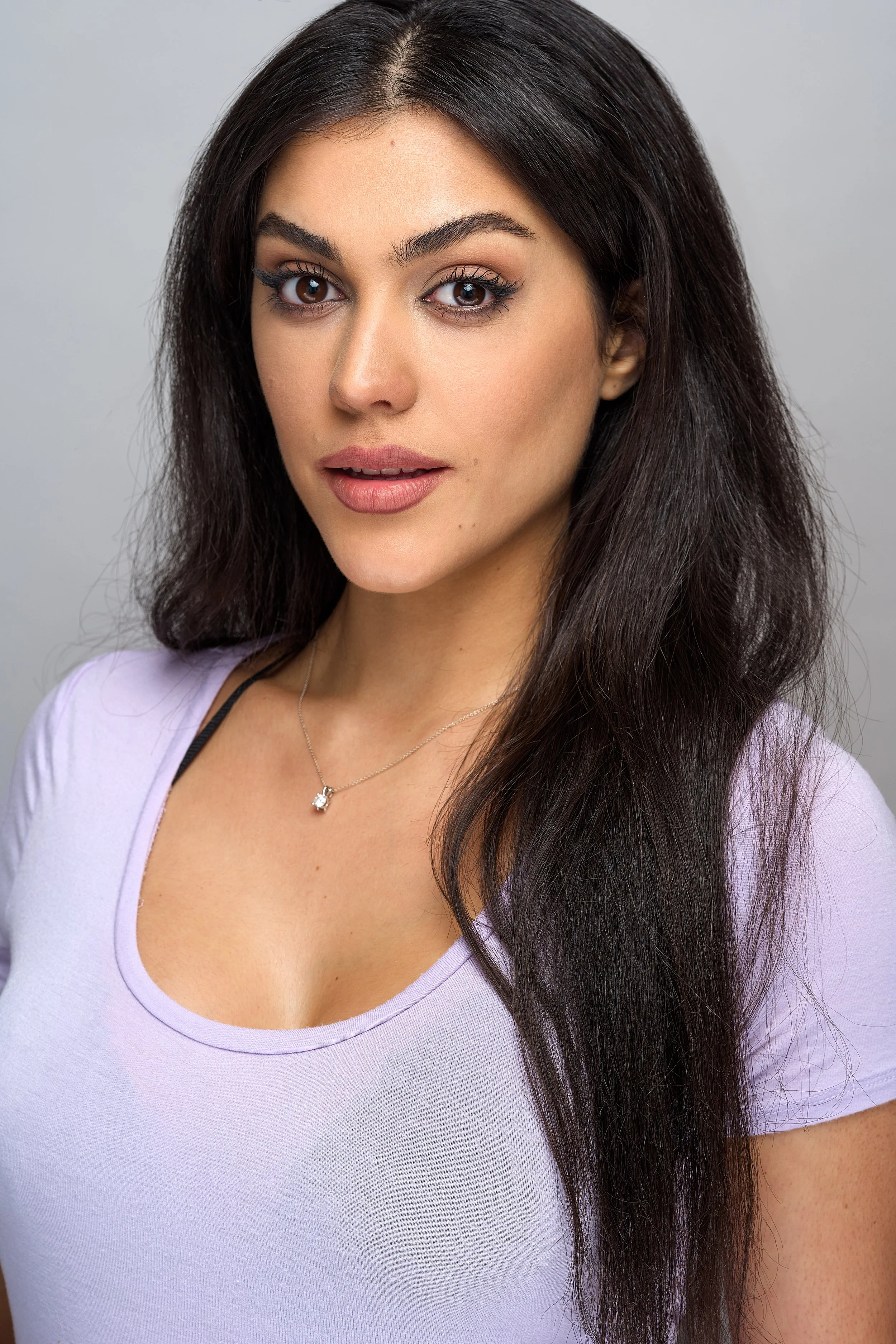Close-up of a young woman with long dark hair, wearing a light purple top and a delicate necklace, against a plain gray background.