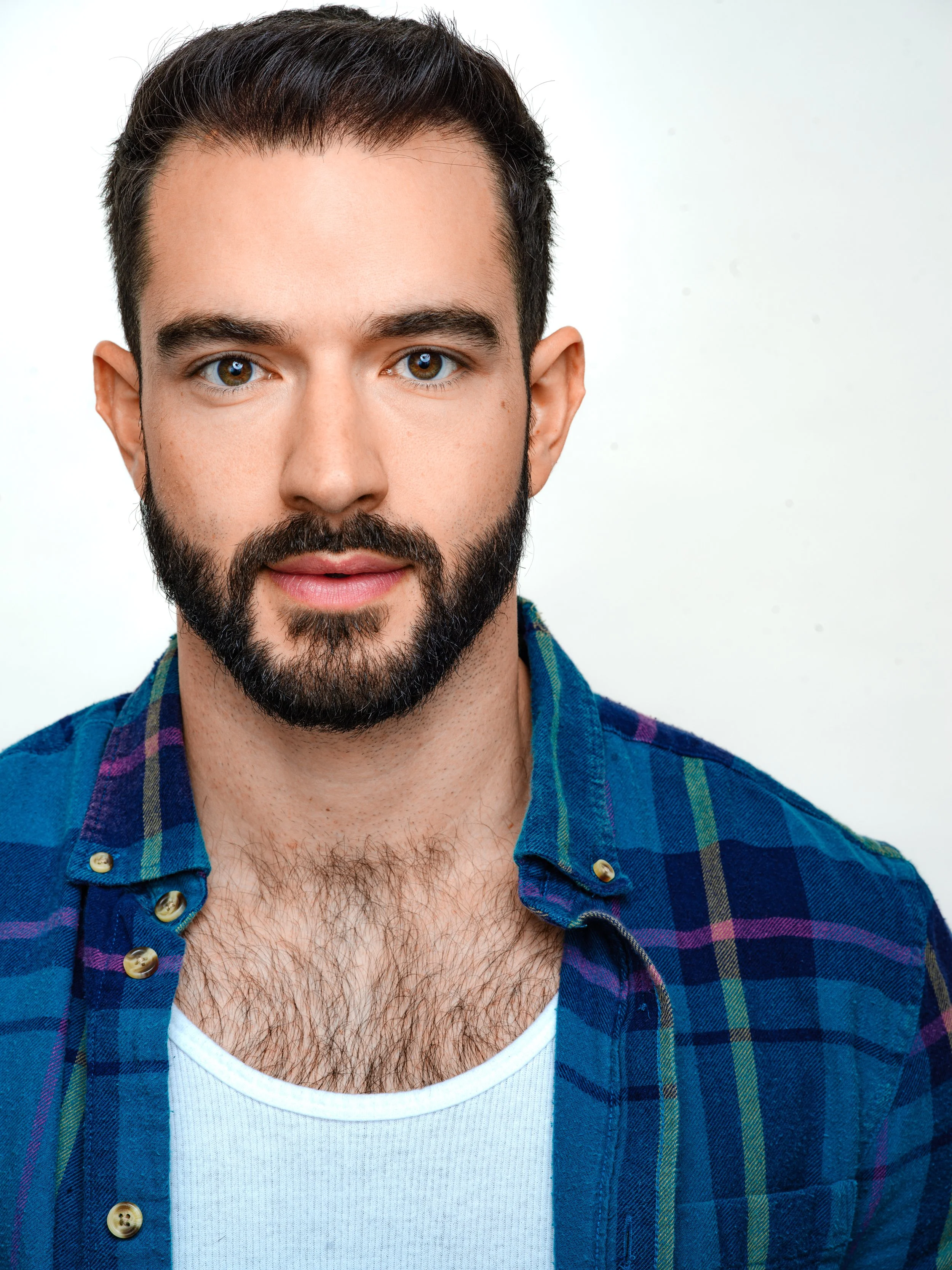 Close-up of a man with dark hair, brown eyes, and a beard wearing a blue plaid shirt over a white t-shirt.