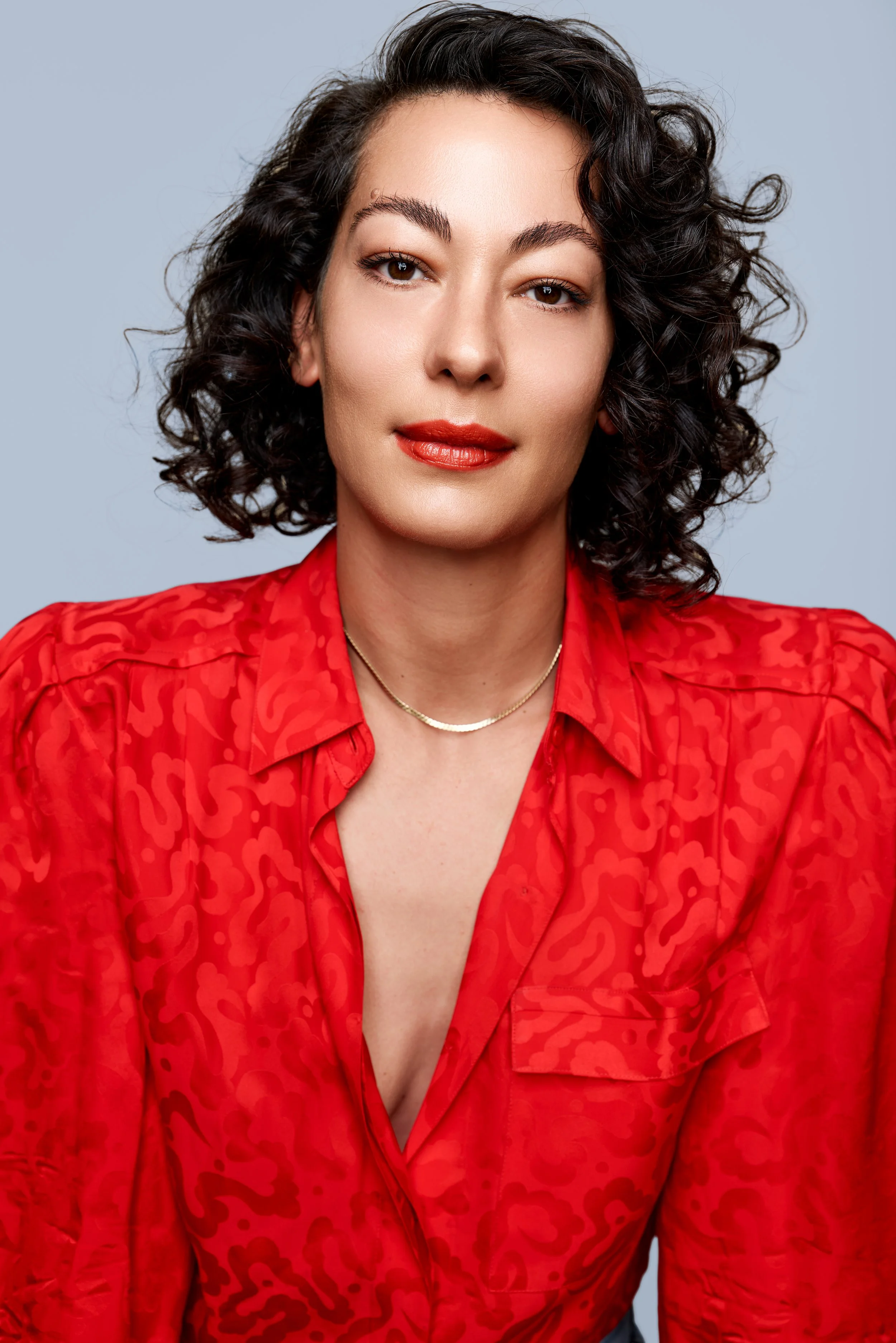 Close-up of a woman with curly dark hair wearing a red patterned blouse and gold necklace, with fair skin and makeup, against a light gray background.