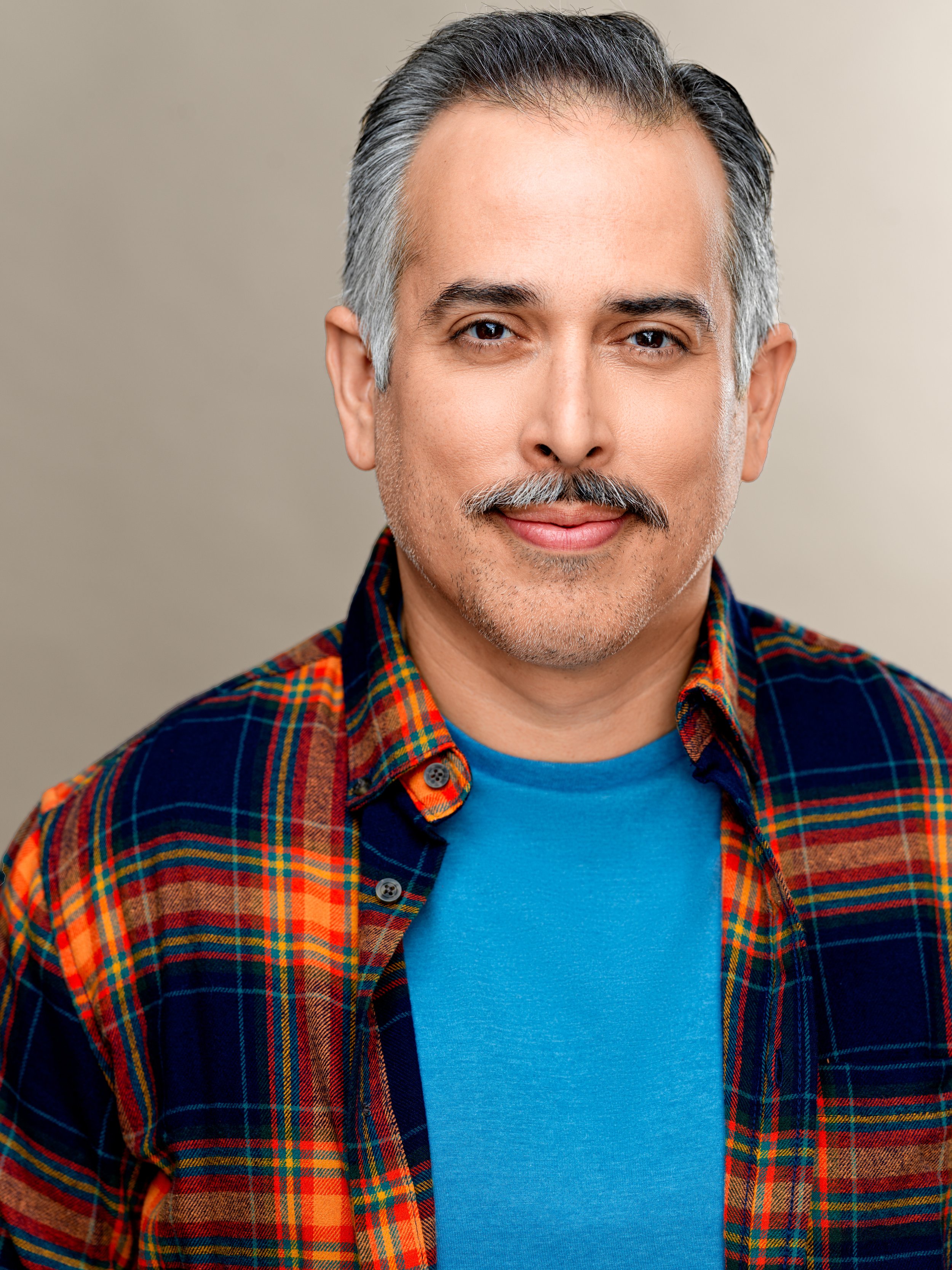 Close-up portrait of a middle-aged man with gray and black hair, a mustache, and a goatee, wearing a blue T-shirt and a colorful plaid shirt, smiling slightly against a neutral background.
