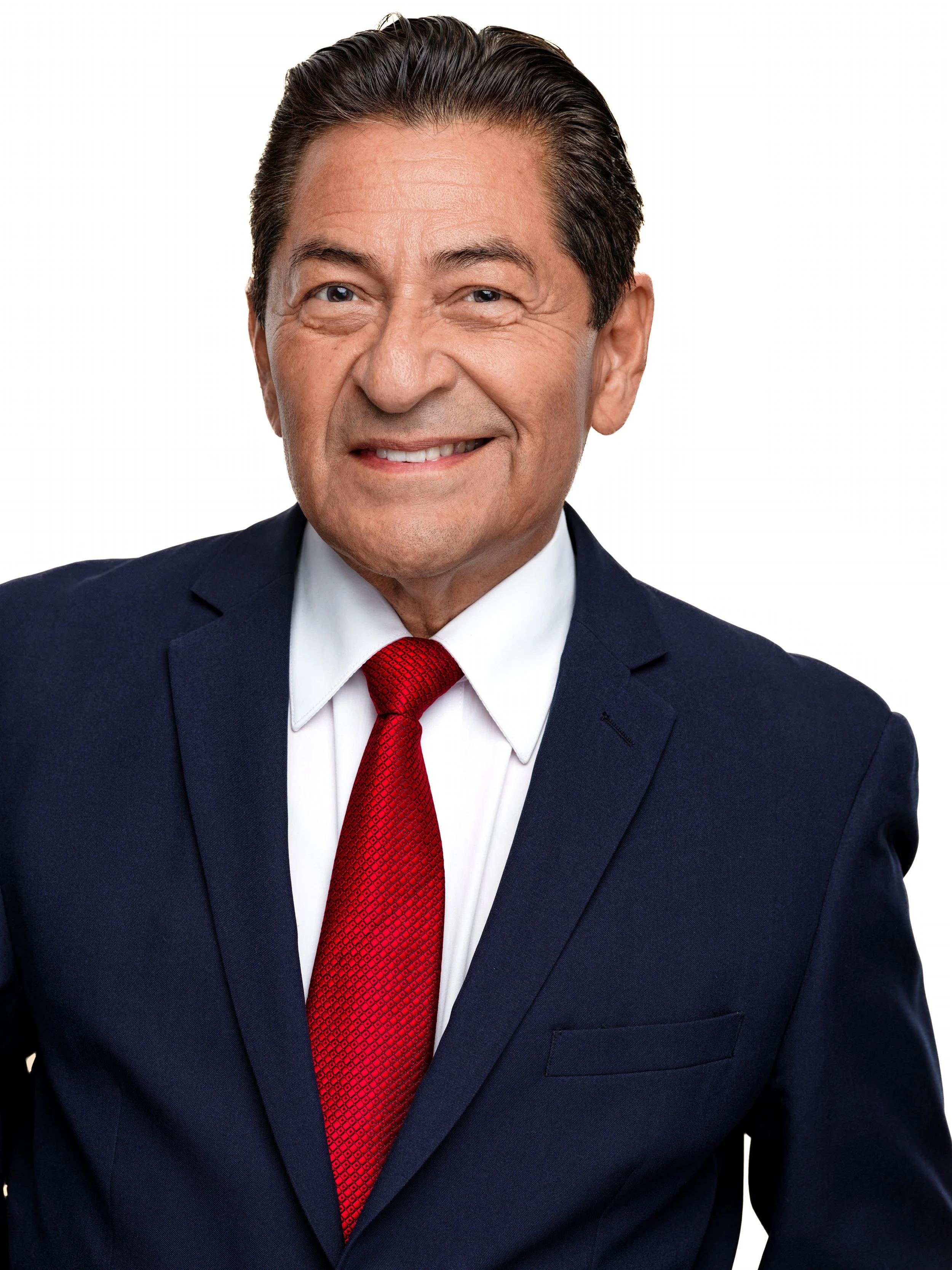 A photograph of a man wearing a navy blue suit with a white shirt and a red tie, smiling and looking directly at the camera.