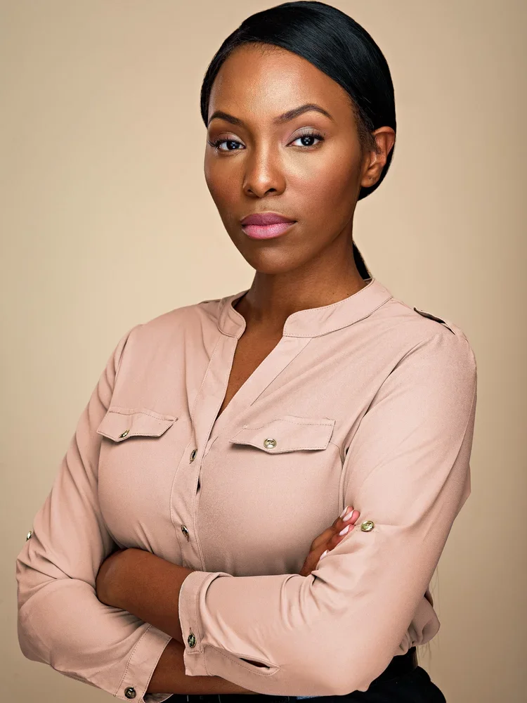 A confident African American woman with black hair and light makeup, wearing a beige button-up blouse with rolled-up sleeves, standing with arms crossed against a neutral background.