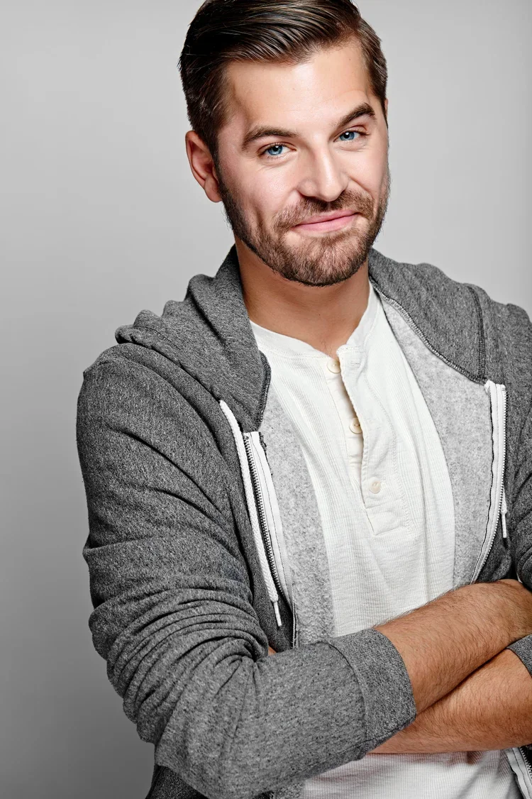 A young man with light skin, blue eyes, and a beard, wearing a gray hoodie and white shirt, smiling confidently with arms crossed against a plain gray background.