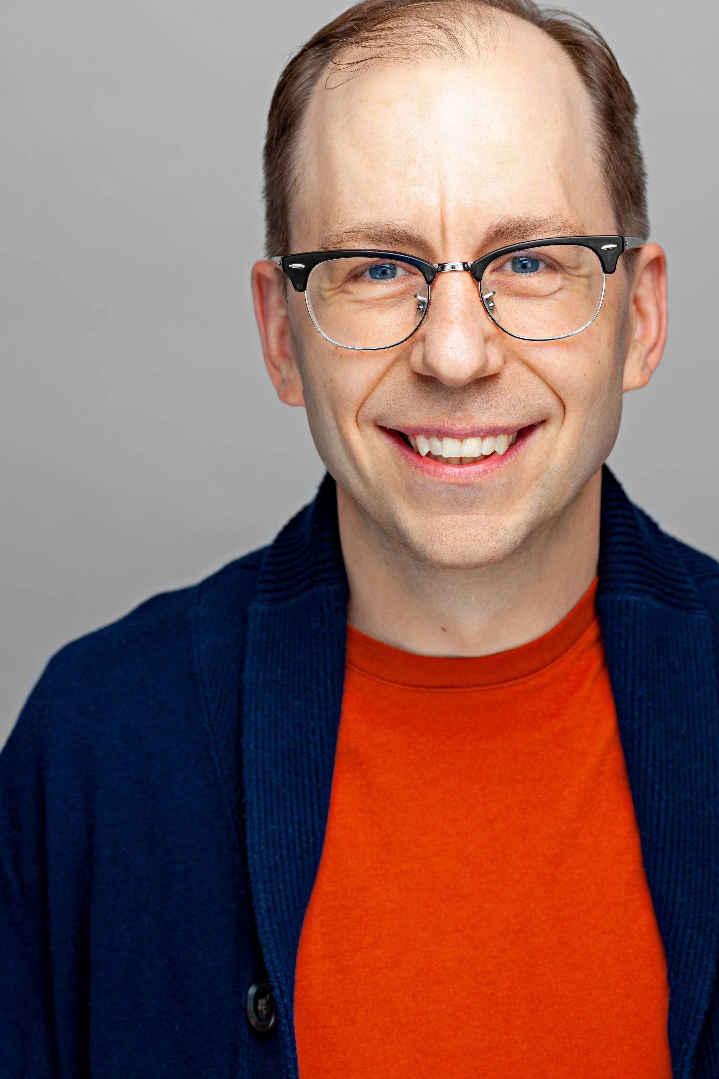 A smiling man with blue eyes and glasses, wearing an orange shirt and a dark blue sweater, against a plain gray background.