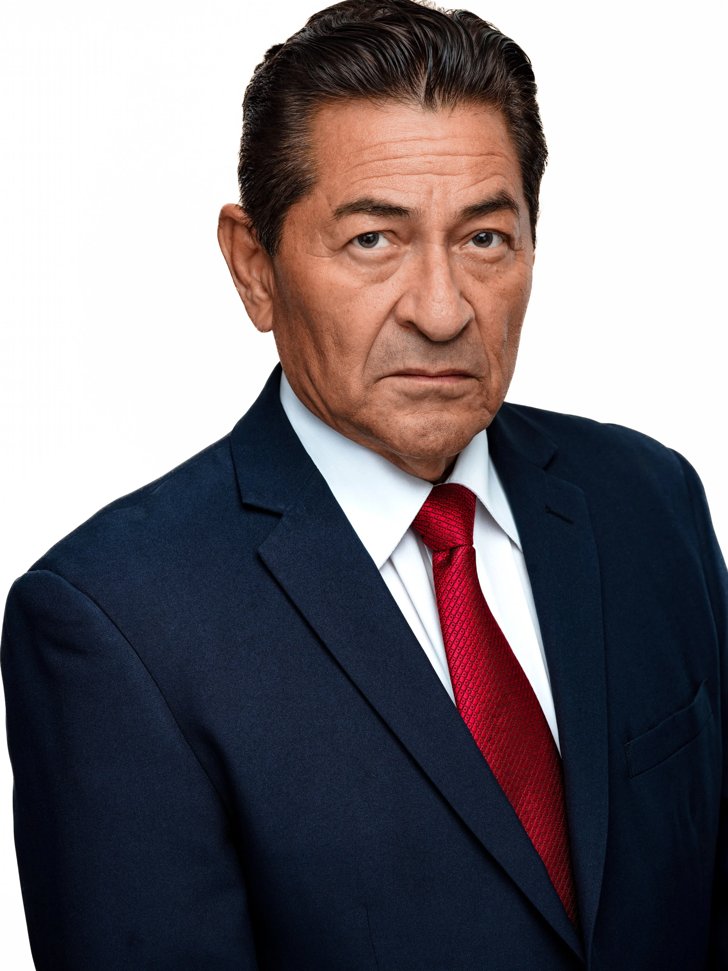 A middle-aged man with dark hair, wearing a navy suit, white dress shirt, and red tie, looking serious with crossed arms against a white background.