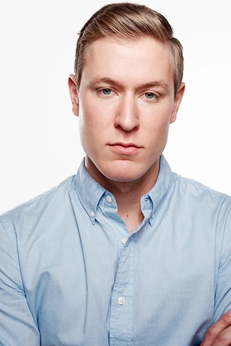A young man with light skin and short blonde hair, wearing a light blue button-up shirt, looking directly at the camera with a neutral expression.