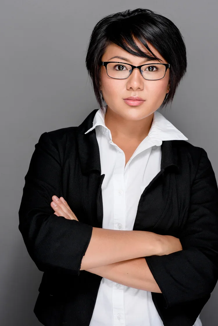 A woman with short black hair, glasses, wearing a white collared shirt and a black blazer, standing with arms crossed against a plain gray background.