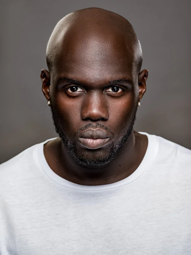 Portrait of a serious-looking Black man with a shaved head and a short beard, wearing earrings and a white T-shirt, against a plain dark background.