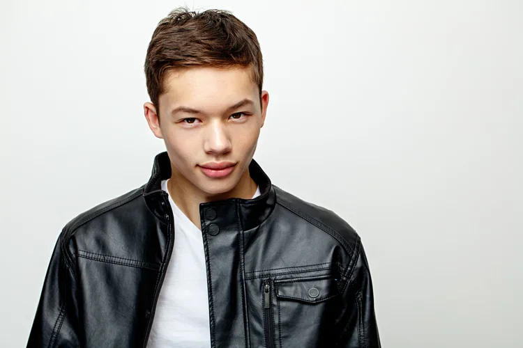Young man with short brown hair wearing a black leather jacket and white T-shirt standing against a plain white background.