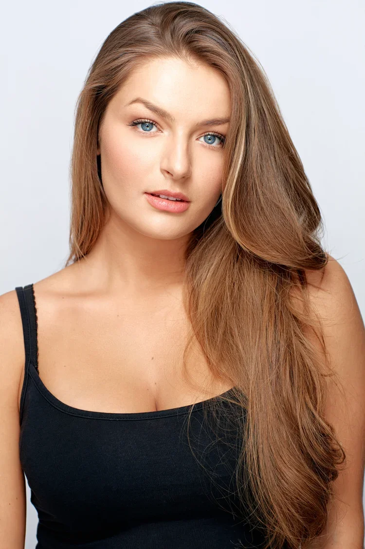 A young woman with long, wavy light brown hair, blue eyes, and a neutral expression, wearing a black tank top, against a plain light background.