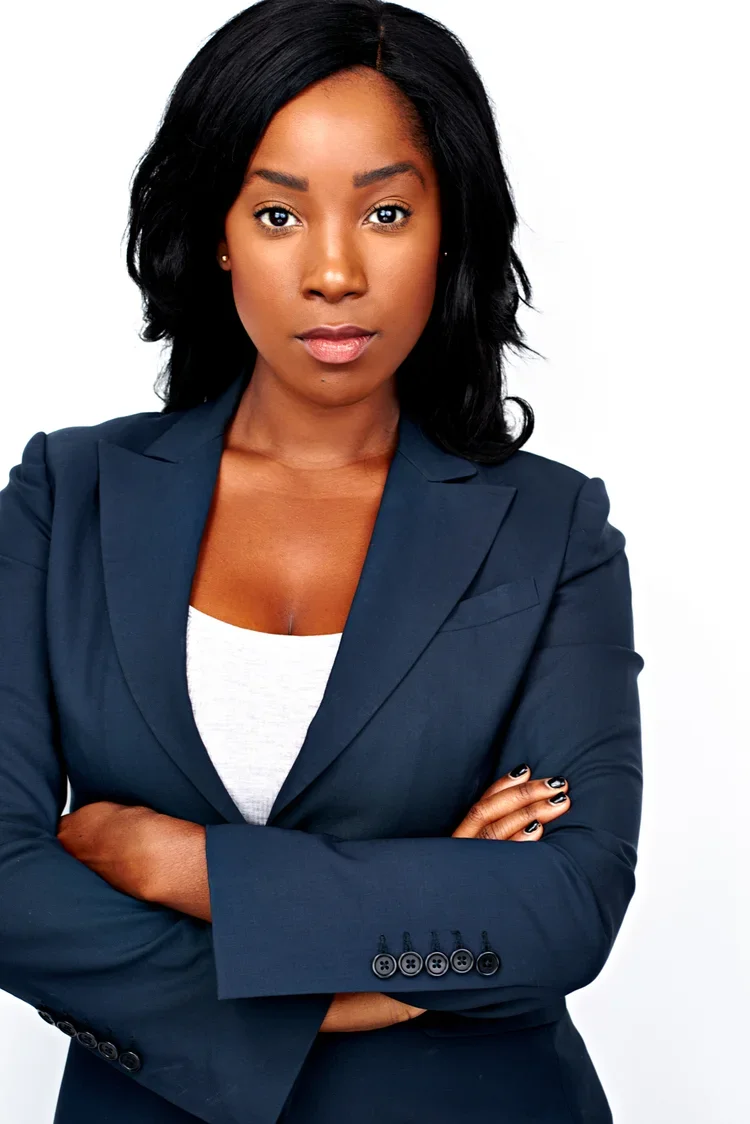 Portrait of a confident African American woman with crossed arms, wearing a navy blazer and white top, standing against a white background.