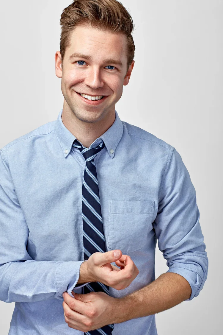 Smiling young man in a light blue shirt and a striped tie, standing against a plain gray background.