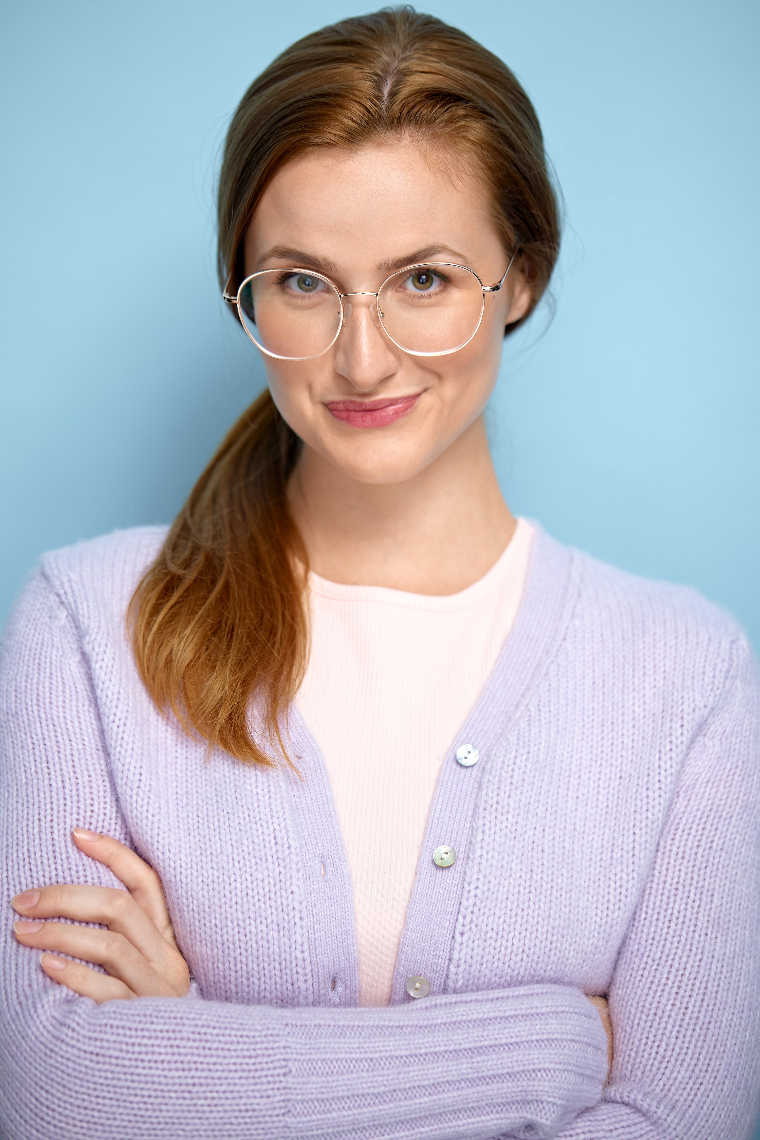 Close-up of a young woman with long red hair, wearing round glasses and a pastel purple cardigan, smiling slightly against a blue background.